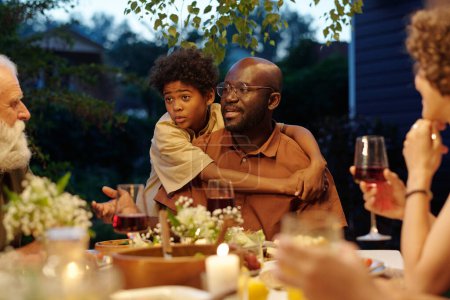 Foto de Lindo niño y su padre mirando al hombre mayor mientras habla con él por la mesa servida con comida casera, bebidas y racimos de flores - Imagen libre de derechos