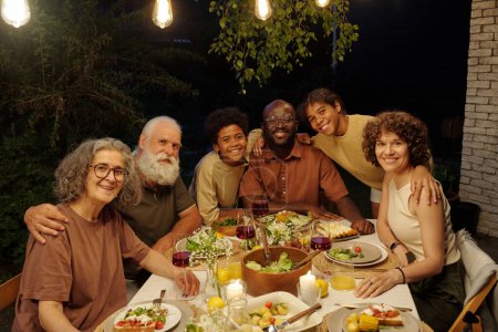 Photo for Cheerful intercultural family of three generations sitting by served table during outdoor dinner in the evening by their country house - Royalty Free Image
