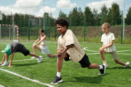 Foto de Grupo de escolares haciendo ejercicio físico sobre hierba verde del campo de fútbol o estadio mientras lo repiten después de un colegial afroamericano - Imagen libre de derechos