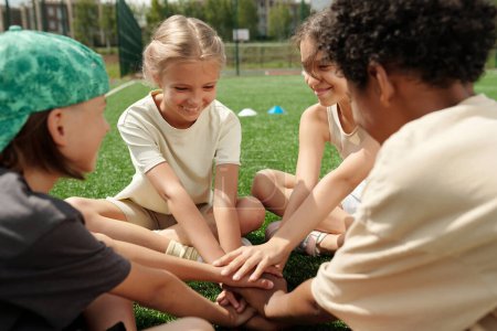 Photo for Cute smiling schoolgirl and her classmates making pile of hands while sitting in front of camera on football field during outdoor lesson - Royalty Free Image