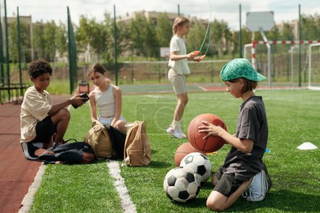 Téléchargez les photos : Vue latérale de l'écolier en vêtements de sport choisir la balle pour jouer au jeu de sport tout en se tenant sur ses genoux sur la pelouse verte du terrain de football - en image libre de droit