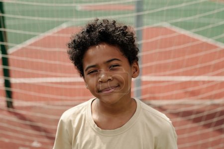 Photo for Happy cute African American schoolboy looking at camera against goal net while standing on sports ground or stadium on sunny day - Royalty Free Image