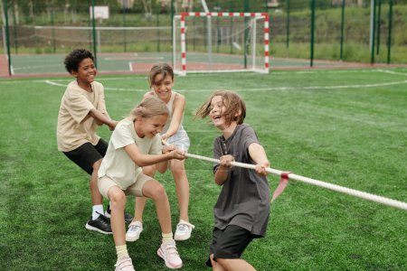 Téléchargez les photos : Des écoliers joyeux en vêtements de sport riant tout en tirant la corde pendant la compétition sportive avec une autre équipe sur le terrain de football vert au stade - en image libre de droit