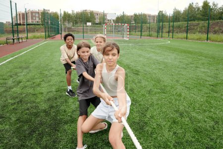 Photo for Group of four strong intercultural classmates pulling rope at lesson of physical training or during sports competition on football field - Royalty Free Image