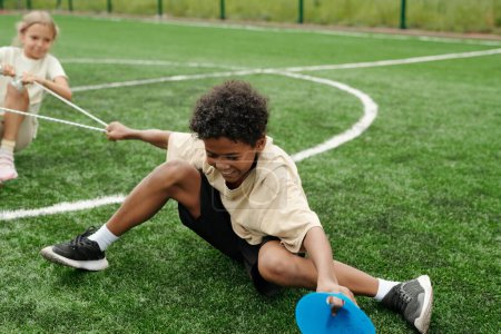 Téléchargez les photos : Joyeux écolier afro-américain tirant la corde et résistant à son camarade de classe lors d'une compétition sportive sur un stade ou un terrain de football - en image libre de droit