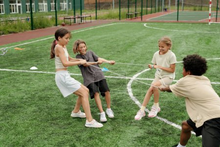 Téléchargez les photos : Quatre écoliers interculturels mignons en vêtements de sport tirant la corde au stade tout en se tenant debout sur le terrain de football vert et l'entraînement avant le match - en image libre de droit