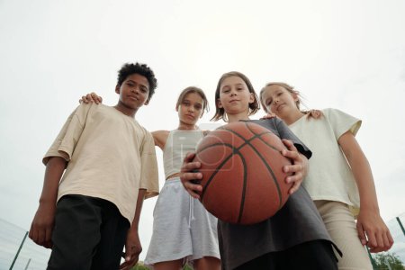 Foto de Bajo ángulo de cuatro chicos y chicas interculturales serios en ropa deportiva mirando a la cámara mientras uno de ellos sostiene la pelota para jugar baloncesto - Imagen libre de derechos
