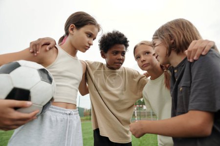 Foto de Equipo de fútbol de cuatro lindos escolares interculturales consultando sobre el juego mientras están de pie frente a la cámara en el descanso después del juego - Imagen libre de derechos
