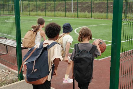 Téléchargez les photos : Vue arrière de quatre écoliers entrant dans un terrain de sport ou un stade avec un terrain de balle de foire tandis que l'un d'eux dribble ball pour jouer au basket-ball - en image libre de droit