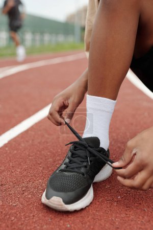 Foto de Céntrate en la pierna del chico afroamericano atando el cordón de la zapatilla antes de correr whil.e de pie en la pista de carreras delante de la cámara en el estadio - Imagen libre de derechos