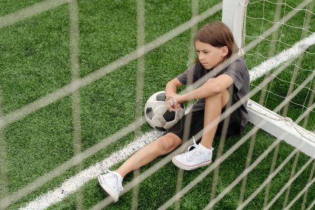 Photo for Sad restful schoolboy in activewear sitting on green football field behind goal net and having rest after hard training before competition - Royalty Free Image