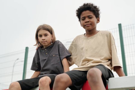 Téléchargez les photos : Deux écoliers interculturels mignons en t-shirts regardant la caméra tout en étant assis sur le filet de but et en ayant du repos entre les entraînements sportifs sur le stade - en image libre de droit