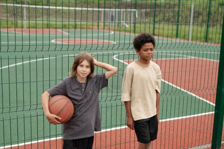 Téléchargez les photos : Deux garçons interculturels reposants en t-shirts debout près d'une clôture métallique verte entourant un grand terrain de sport pour jouer au football ou au basket-ball - en image libre de droit