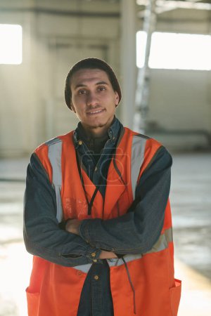 Photo for Happy young engineer or foreman in protective fluorescent vest looking at camera with his arms crossed by chest while standing in workshop - Royalty Free Image