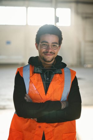 Téléchargez les photos : Jeune ingénieur multiethnique masculin en lunettes et vêtements de travail regardant la caméra alors qu'il se tenait dans un entrepôt avec les bras croisés par la poitrine - en image libre de droit