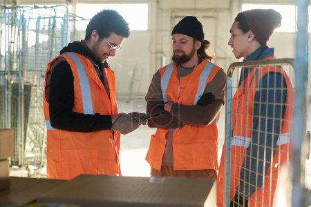 Photo for Three young male workers of warehouse in reflective jackets discussing list of delivered goods while standing by cart with stack of boxes - Royalty Free Image