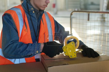 Photo for Cropped shot of young warehouse worker in safety vest and gloves packing metallic spare parts into boxes and sealing them with cellotape - Royalty Free Image
