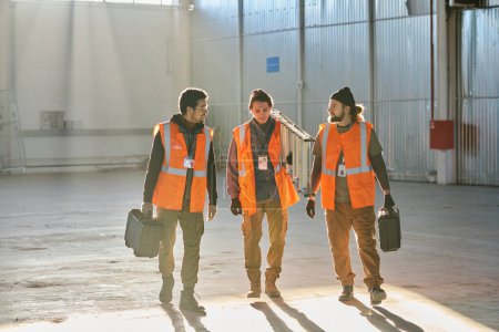 Téléchargez les photos : Trois jeunes ingénieurs interculturels en vêtements de travail se déplaçant le long de l'entrepôt tandis que deux d'entre eux avec des boîtes à outils regardant collègue avec escabeau - en image libre de droit