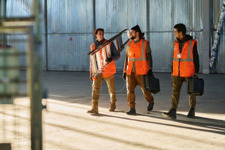 Téléchargez les photos : Trois heureux ingénieurs d'entrepôt avec escabeau et boîtes à outils se regardant et bavardant tout en marchant vers l'avant - en image libre de droit