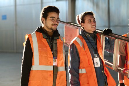 Téléchargez les photos : Deux jeunes travailleurs d'entrepôt multiethniques en gilets fluorescents de protection debout devant la caméra en atelier et regardant vers l'avenir - en image libre de droit