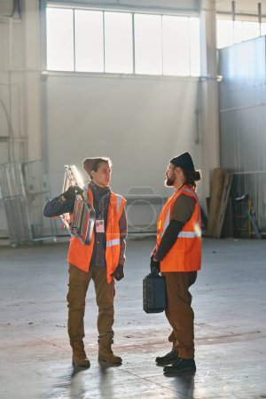 Téléchargez les photos : Deux jeunes ingénieurs en vestes réfléchissantes protectrices discutent de quelque chose lors d'une réunion de travail tandis que l'un d'eux écoute un collègue - en image libre de droit