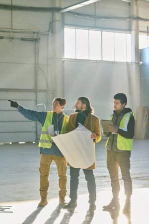 Téléchargez les photos : Jeune ingénieur confiant pointant vers le mur de l'entrepôt lors de la discussion des points de travail et du plan de construction détenu par l'entrepreneur - en image libre de droit
