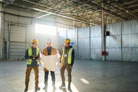 Téléchargez les photos : Trois ingénieurs portant un casque de protection discutent du plan avec le plan du bâtiment tout en se tenant dans un entrepôt contre la lumière du soleil qui passe par la fenêtre - en image libre de droit