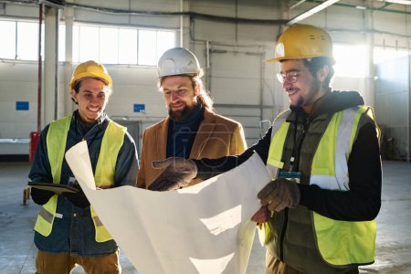Téléchargez les photos : Jeune ingénieur souriant pointant du doigt le plan de construction dans le plan directeur tout en l'expliquant à l'entrepreneur et à son collègue lors d'une réunion dans un entrepôt - en image libre de droit