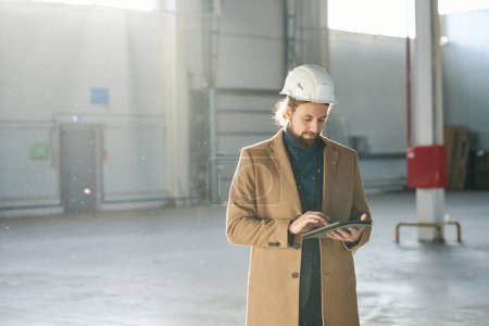 Photo for Young bearded builder or solopreneur in hardhat scrolling in tablet while standing in front of camera in spacious sunlit warehouse and networking - Royalty Free Image
