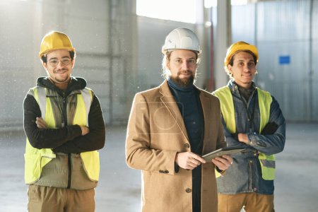 Téléchargez les photos : Heureux jeune contremaître en manteau beige et deux ingénieurs multidisciplinaires en casques de protection et vestes réfléchissantes de sécurité debout devant la caméra - en image libre de droit