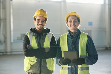 Foto de Dos jóvenes ingenieros multiétnicos felices en sombreros y chaquetas reflectantes de pie delante de la cámara en el almacén de la planta industrial o fábrica - Imagen libre de derechos