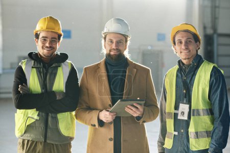 Téléchargez les photos : Trois jeunes ingénieurs interculturels heureux dans des casques de protection regardant la caméra avec des sourires tandis que deux hommes en gilets de sécurité entourant contremaître - en image libre de droit
