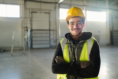 Téléchargez les photos : Jeune ingénieur joyeux en casque de sécurité, gilet de protection et lunettes regardant la caméra avec le sourire tout en restant debout dans un entrepôt spacieux - en image libre de droit