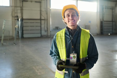 Téléchargez les photos : Jeune ingénieur ou constructeur prospère en vêtements de travail protecteurs regardant la caméra avec le sourire tout en restant debout dans l'entrepôt et en utilisant une tablette - en image libre de droit