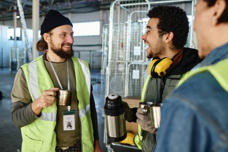 Téléchargez les photos : Jeune ingénieur joyeux avec tasse de thermos riant tout en parlant à un collègue avec une tasse métallique avec du thé chaud pendant la pause dans l'entrepôt - en image libre de droit