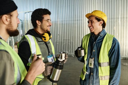 Photo for Focus on two young cheerful multiethnic engineers in reflective vests looking at one another during tea break while holding metallic mugs - Royalty Free Image
