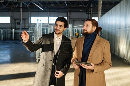 Photo for Young confident owner of industrial factory showing new equipment to male colleague in formalwear holding tablet and looking forwards - Royalty Free Image