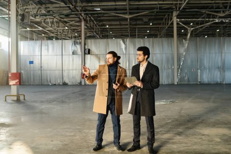 Téléchargez les photos : Long shot de deux jeunes entrepreneurs interculturels en vêtements de formalerie regardant autour de la zone pour de nouveaux bureaux tout en se tenant dans un entrepôt spacieux - en image libre de droit
