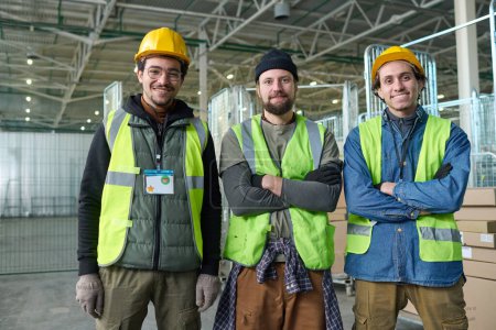 Téléchargez les photos : Groupe de jeunes travailleurs interculturels heureux de l'entrepôt ou de l'usine dans des casques de sécurité et vestes fluorescentes debout devant la caméra - en image libre de droit