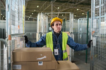 Téléchargez les photos : Jeune travailleur de l'entrepôt dans le casque de sécurité et gilet réfléchissant poussant grand chariot métallique avec des piles de boîtes tout en avançant vers l'avant - en image libre de droit