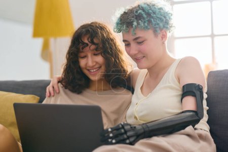 Photo for Happy young woman with disability and her girlfriend watching online video while sitting on couch at home and looking at screen of laptop - Royalty Free Image