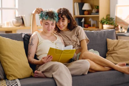 Foto de Joven morena sonriente sentada en el sofá junto a su novia con discapacidad leyendo libro mientras ambos pasan tiempo en el entorno doméstico - Imagen libre de derechos