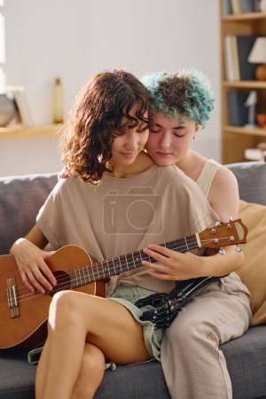 Foto de Amputada joven femenina con mano mioeléctrica abrazando a su novia con guitarra acústica mientras ambos están sentados en un cómodo sofá en casa - Imagen libre de derechos