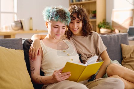 Foto de Mujer joven con la mano mioeléctrica y su novia mirando a la página de libro abierto en la cubierta amarilla, mientras que ambos sentados en el sofá en el ocio - Imagen libre de derechos