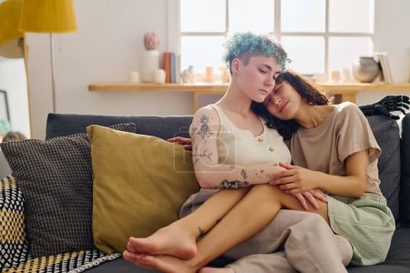 Young serene lesbian couple in casualwear enjoying rest on comfortable couch with cushions against window and interior of living room