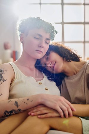 Téléchargez les photos : Deux jeunes amies interculturelles sereines gardant les yeux fermés tout en se relaxant sur le canapé devant la caméra dans le salon de leur appartement - en image libre de droit