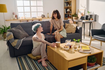 Photo for Two happy young girlfriends in casualwear looking at one another during breakfast while sitting in living room, having tea and chatting - Royalty Free Image