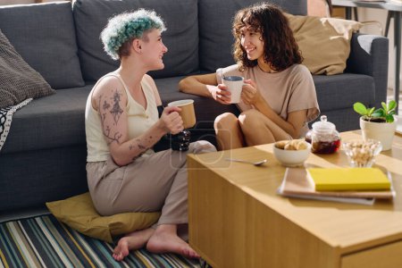 Photo for Happy young woman with myoelectric hand and her girlfriend sitting on the floor between couch and table, chatting and having tea - Royalty Free Image