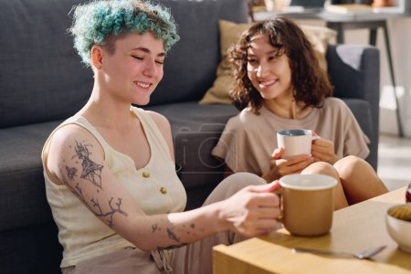 Photo for Young cheerful woman with cup of tea or coffee and her brunette girlfriend sitting by table against couch and having chat while enjoying home rest - Royalty Free Image
