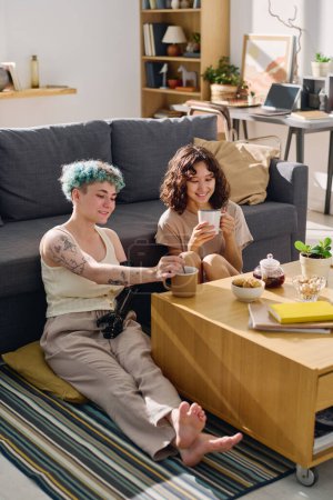 Téléchargez les photos : Deux jeunes femmes heureuses assises par terre sur le canapé et prenant le thé pour le petit déjeuner tandis que l'une d'elles gardait la main myoélectrique sur le ventre - en image libre de droit